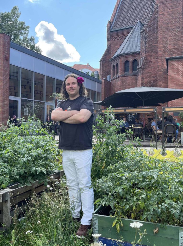 Person mit langem Haar, Blume hinter dem Ohr, Arme verschränkt, steht in einem Gartencafé.