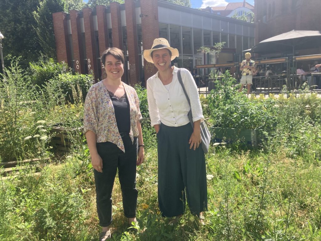 Zwei Frauen lächeln in einem sonnigen Garten mit hohen Pflanzen und Backsteinbauten im Hintergrund.