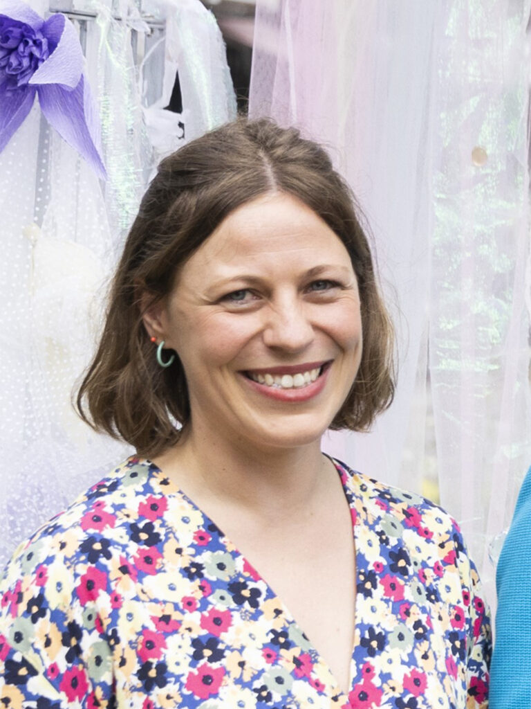 Lächelnde Frau mit schulterlangen braunen Haaren in einem geblümten Kleid, die vor einem pastellfarbenen Stoff steht.