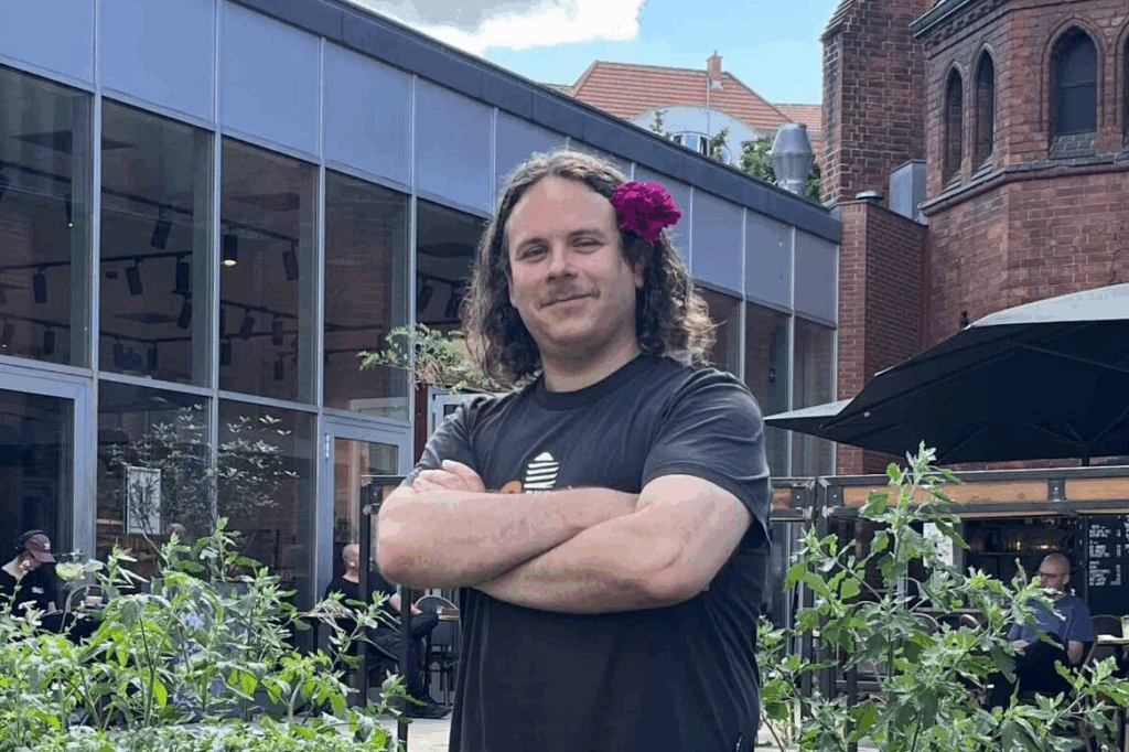 Eine Person mit langen Haaren und einer Blume im Ohr steht mit verschränkten Armen in einem Garten in der Nähe moderner Gebäude.