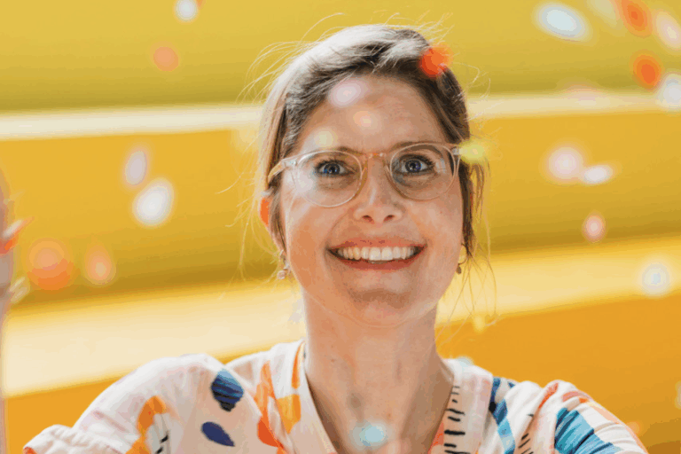 Lächelnde Frau mit Brille und einem bunten Hemd mit Konfetti auf gelbem Hintergrund.